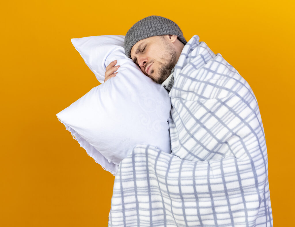sleepy young blonde ill slavic man wearing winter hat and scarf wrapped in plaid holds and puts head on pillow isolated on orange background with copy space